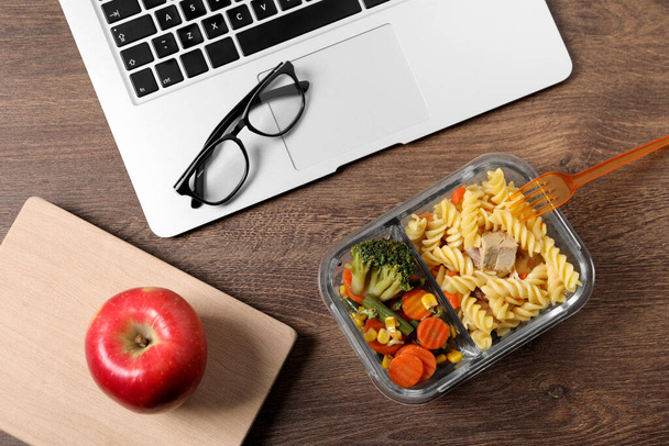 Container of tasty food, laptop, apple, book and glasses on wooden table, flat lay. Business lunch - Фото, изображение