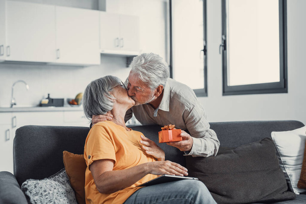 Smiling senior husband making surprise giving gift box to amazed wife, happy aged woman get unexpected present from spouse spending romantic weekend or celebrating anniversary together - 写真・画像