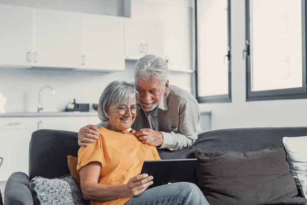 Glückliches älteres Paar, das zu Hause gemeinsam ein Tablet benutzt, aufgeregter älterer Mann und Frau, die auf den Bildschirm des Mobilgeräts schauen, einkaufen oder online chatten, auf der gemütlichen Couch im Wohnzimmer sitzen - Foto, Bild