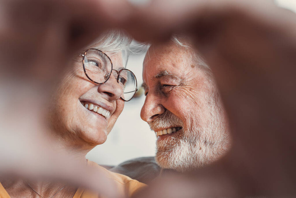 Close up portrait happy sincere middle aged elderly retired family couple making heart gesture with fingers, showing love or demonstrating sincere feelings together indoors, looking at camera. - Foto, Imagen