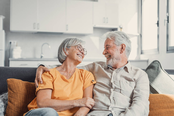 Счастливая смеющаяся пожилая супружеская пара разговаривает, смеется, стоя в интерьере дома вместе, обнимаясь с любовью, наслаждаясь близкими отношениями, доверием, поддержкой, заботой, ощущая радость, нежность - Фото, изображение