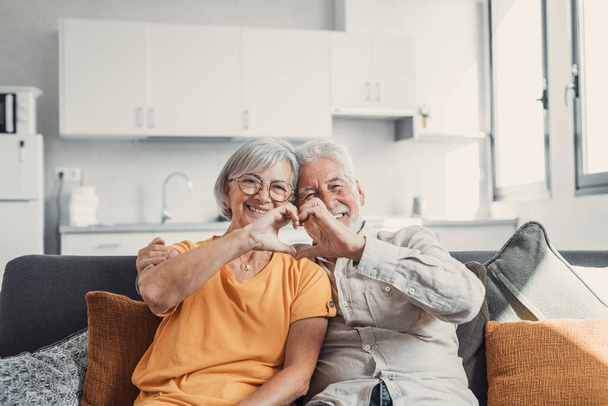Close up portrait happy sincere middle aged elderly retired family couple making heart gesture with fingers, showing love or demonstrating sincere feelings together indoors, looking at camera. - Photo, Image