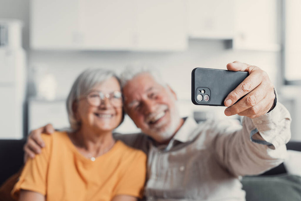 携帯電話で自分撮りをする幸せな老夫婦、笑顔のシニア成熟した配偶者中年の妻と引退した夫がスマートフォンのカメラで自画像を作る携帯電話を持って、モバイルディスプレイに焦点を当てて - 写真・画像