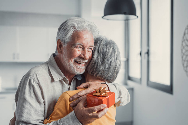 Caring smiling mature elderly woman giving wrapped gift box to smiling old 80s husband, congratulating with birthday or wedding anniversary. Joyful senior man feeling excited getting present from wife - Foto, Imagem