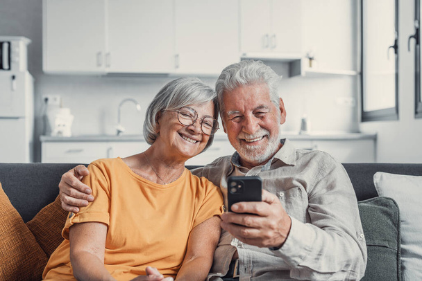 Šťastná dospělá rodina, manželka a manžel používají doma telefon a usmívají se muž a žena středního věku, kteří sedí na pohovce, používají aplikace pro mobilní zařízení, sledují video v sociální síti, surfování po internetu - Fotografie, Obrázek