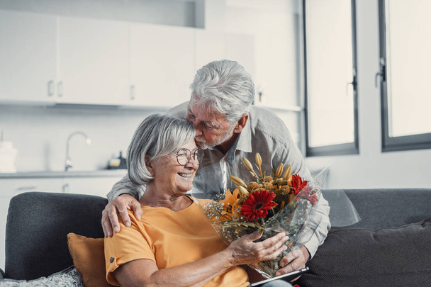 Vanha mies antaa kukkia vaimolleen istuu sohvalla kotona San Ystävänpäivä. Eläkeläiset nauttivat yllätyksestä yhdessä. Rakkaudessa ihmiset pitävät hauskaa. - Valokuva, kuva