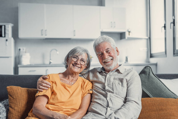 Headshot portrait of smiling elderly 60s husband and wife sit relax on couch hugging cuddling, happy mature old couple rest on sofa in living room embrace look at camera show love and care - Photo, Image