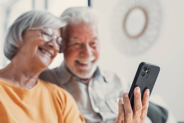 Szczęśliwa dojrzała Rodzina, żona i mąż za pomocą telefonu razem w domu, uśmiechając się w średnim wieku mężczyzna i kobieta siedzi na kanapie, za pomocą aplikacji urządzenia mobilnego, oglądanie wideo w sieci społecznej, surfowanie po Internecie - Zdjęcie, obraz