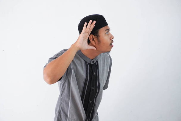 灰色のイスラム教徒の服を着た若いアジア系イスラム教徒の男、白い背景に隔離されたゴシップを聞こうと耳の近くの手 - 写真・画像