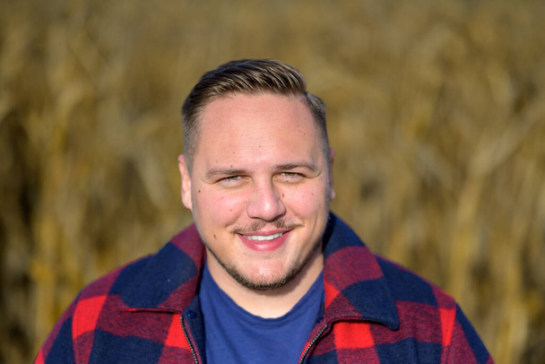 Фронтальный портрет молодого человека с дружелюбной улыбкой в красно-синей куртке лесоруба и синей футболке на осеннем кукурузном поле на заднем плане - Фото, изображение