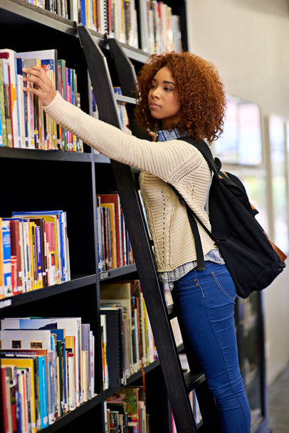 Time to do some research. a young woman reaching for a book from a bookshelf in a university library - Foto, afbeelding