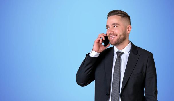 Happy businessman calling on the phone, portrait in black formal suit on copy space blue background. Concept of business network, online communication and conference - Фото, зображення