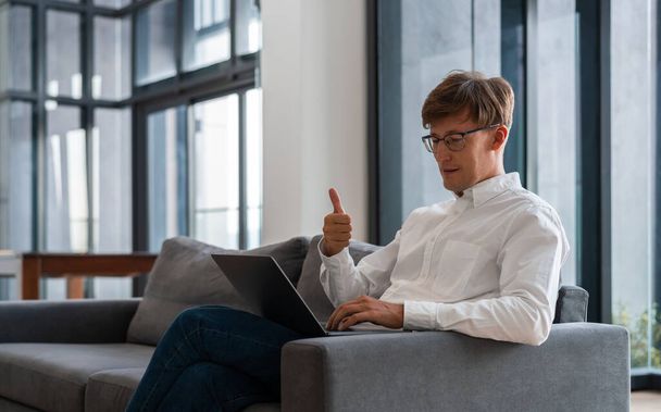 Young businessman with thumb up looking at laptop, video conference and distance work. Man sitting on sofa at home. Concept of online network and communication - Fotoğraf, Görsel