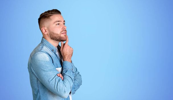 Side portrait of bearded young man in casual clothes standing over blue background. Concept of planning and choice. Mock up - Zdjęcie, obraz