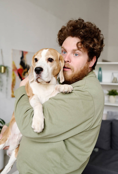 Bearded overweight guy holding his cute pet on arms with scared expression standing in room - Φωτογραφία, εικόνα