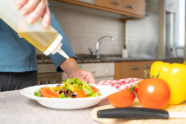 Close up of woman putting olive oil on a fresh salad of tomatoes, lettuce and carrots in the kitchen at home. Healthy and easy to make food concept. High quality photo - Fotó, kép