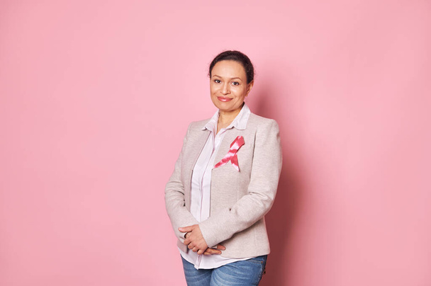 Positive middle-aged Latin American woman, wearing a pink satin ribbon, showing her support for Cancer patients and survivors. World Breast Cancer Day. Awareness campaign. Pink month. October first - Фото, изображение