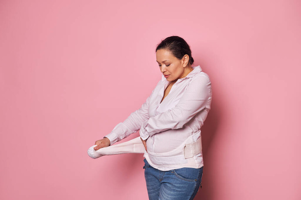 Pregnant woman putting on supporting bandage to reduce backache during pregnancy, isolated over pink background with copy advertising space. Orthopedic abdominal support belt concept. Pregnancy. - Photo, Image