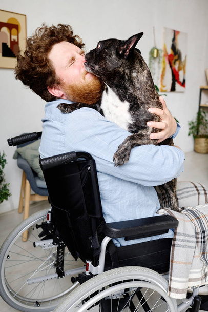 Young beaarded guy in wheelchair closing his eyes while his pet kissing him in face - Фото, зображення