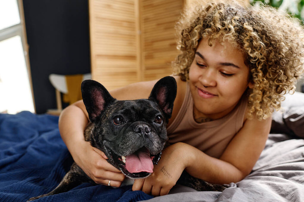 African young girl with curly hair embracing her french bulldog during rest time in bed - Foto, imagen