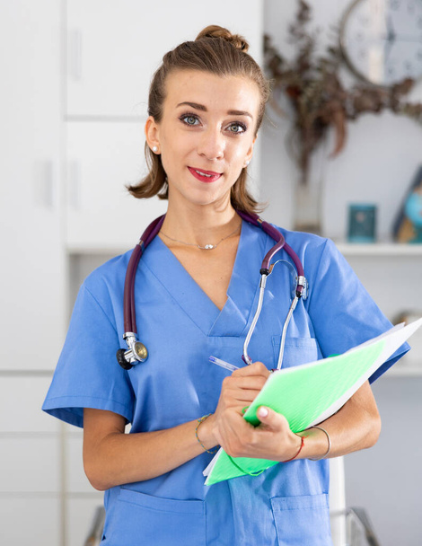 Portrait of smiling female doctor in surgical scrubs holding folder, standing in her office. - 写真・画像