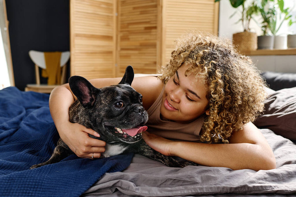 Portrait of happy smiling woman with her cute french bulldog lying on bed at morning - Zdjęcie, obraz