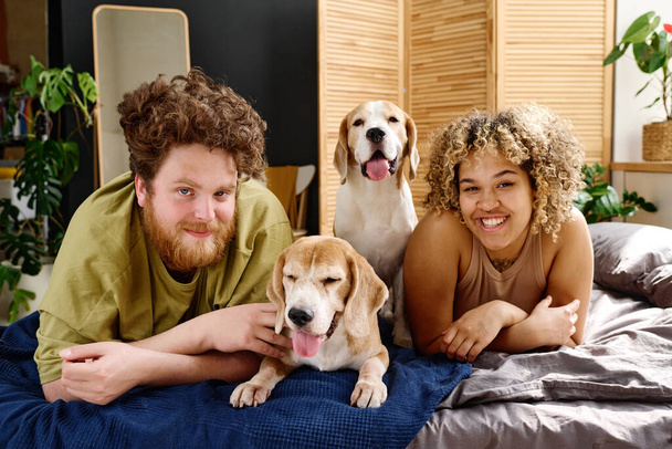 Portrait de jeune couple multiethnique souriant à la caméra allongé sur le lit dans la chambre avec leur animal de compagnie - Photo, image