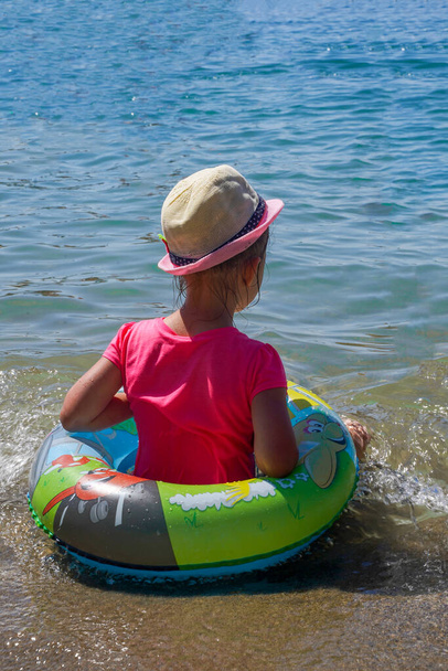 ビーチで円の中の女の子、リアビュー。ピンクのTシャツと藁帽子の子供が海の海岸に座って波を見ています。海の休暇の概念。夏気分. - 写真・画像