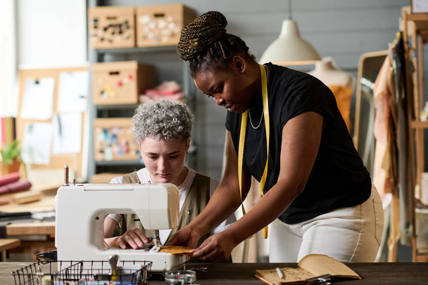 Joven costurera afroamericana ayudando a su colega sentada frente a la máquina de coser eléctrica mientras trabaja sobre una nueva colección - Foto, imagen