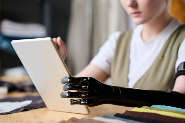 Musta myoelectric käsivarsi proteesi ompelijatar käyttäen tabletti istuessaan työpaikalla ja etsivät aitoja ideoita Internetissä - Valokuva, kuva