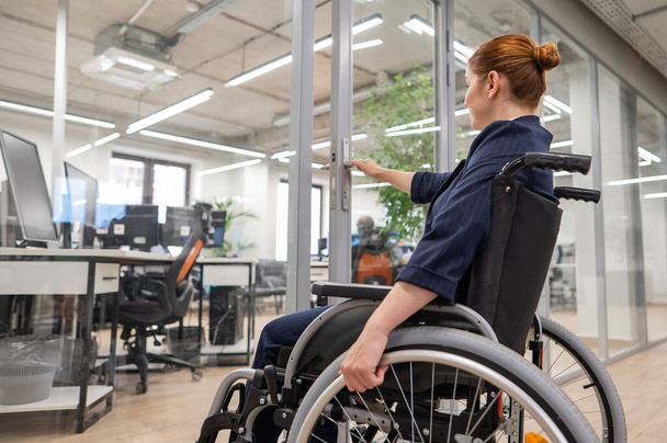 Red-haired caucasian woman in a wheelchair trying to open the door in the office - Fotoğraf, Görsel