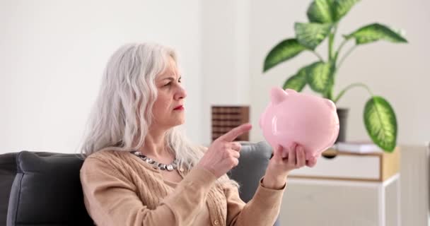 Žádné úspory na penzi a frustrovaná starší žena držící prázdné prasátko. Senior důchodce sedí doma v depresi bez peněz - Záběry, video