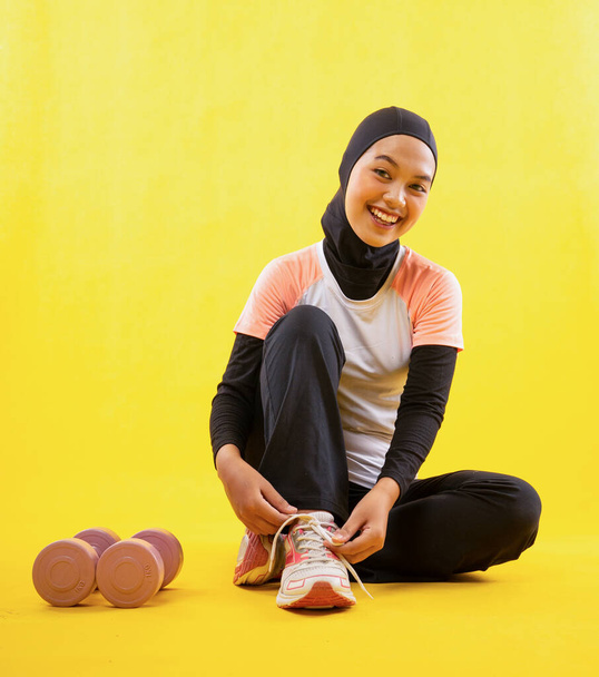 laughing Muslim woman wearing sportswear sit fixing shoelaces on yellow background - Фото, зображення