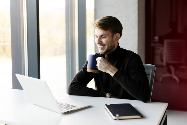 Χαμογελώντας hipster τύπος κρατώντας κύπελλο στο χέρι του, κάθεται στο γραφείο με φορητό υπολογιστή. Ελκυστικός άνθρωπος που εργάζεται στο γραφείο, έννοια του διαλείμματος στην εργασία - Φωτογραφία, εικόνα