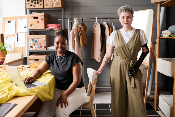 Zwei junge erfolgreiche Schneiderinnen oder Modedesignerinnen blicken in die Kamera, während sie an neuen Projekten oder saisonalen Kollektionen arbeiten - Foto, Bild