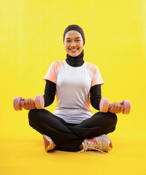 Muslim woman wearing sportswear sit cross-legged exercising using dumbbells on yellow background - Photo, Image