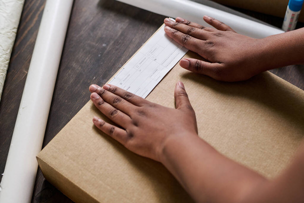 Manos de joven mujer afroamericana poniendo papel con la dirección del cliente en la parte superior de la caja embalada con mercancías ordenadas - Foto, Imagen