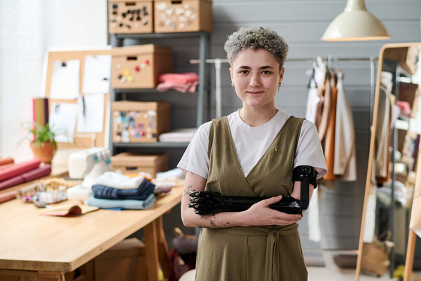 Jonge succesvolle vrouwelijke kleermaker met gedeeltelijke arm staande tegen haar werkplek met tafel, etalagepop, textiel en andere benodigdheden - Foto, afbeelding