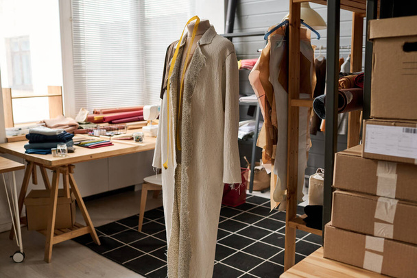 Незаконченное пальто пастельного цвета висит на манекене у стола с припасами внутри просторной мастерской или студии модного дизайна - Фото, изображение