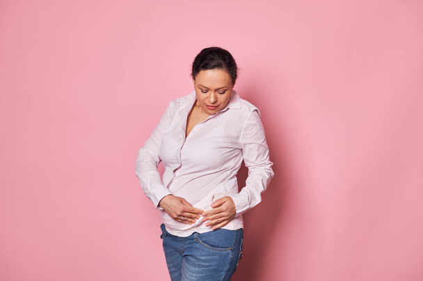 Впевнена темноволоса вагітна жінка середнього віку в повсякденній білій футболці та блакитних джинсах, одягаючи ортопедичні пов'язки для підтримки живота, ізольовані на рожевому тлі. Концепція жіночого здоров'я
 - Фото, зображення