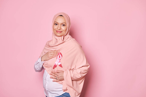 Mujer embarazada musulmana positiva con la cabeza cubierta de hiyab rosa, cogida de la mano alrededor de una cinta de satén rosa, mostrando su apoyo y empatía por los sobrevivientes de cáncer y los pacientes, aislados en el fondo rosa - Foto, Imagen