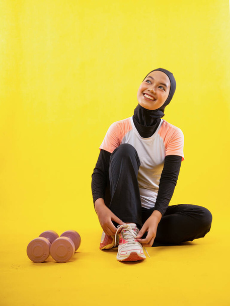 cheerful Muslim woman wearing sportswear sits fixing shoelaces on a yellow background. - Фото, изображение