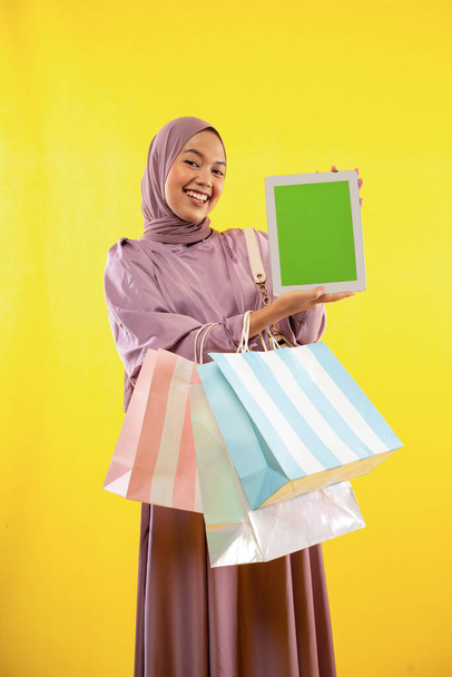 muslim woman in pink hijab dress with shopping bag showing pad screen on yellow background - Fotoğraf, Görsel