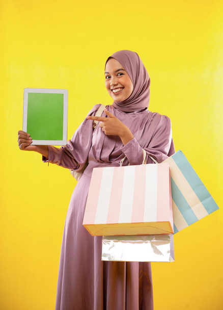 mujer con vestido de hijab muestra la pantalla de la tableta con el dedo apuntando mientras que las compras en el fondo amarillo - Foto, Imagen