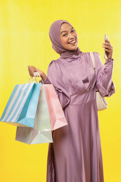 smiling Asian woman in pink hijab dress holding shopping bag and cellphone on yellow background - Photo, image