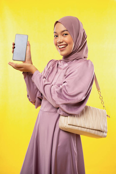 attractive asian muslimah woman shows blank phone screen on yellow background - Fotó, kép