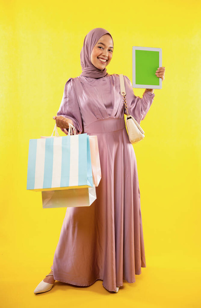 smiling Asian woman in pink hijab dress holding shopping bag and tablet on yellow background - Foto, Bild