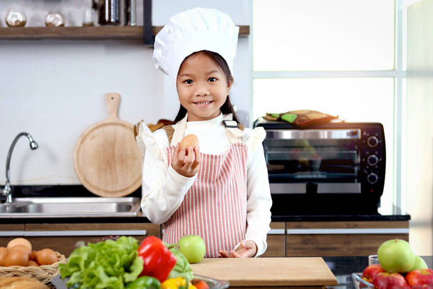 Ritratto di ragazza asiatica sorridente bambino che indossa grembiule e cappello da chef in piedi al bancone della cucina con verdure fresche colorate ingredienti alimentari insalata, bambino cuoco carino preparare la cucina cibo sano. - Foto, immagini