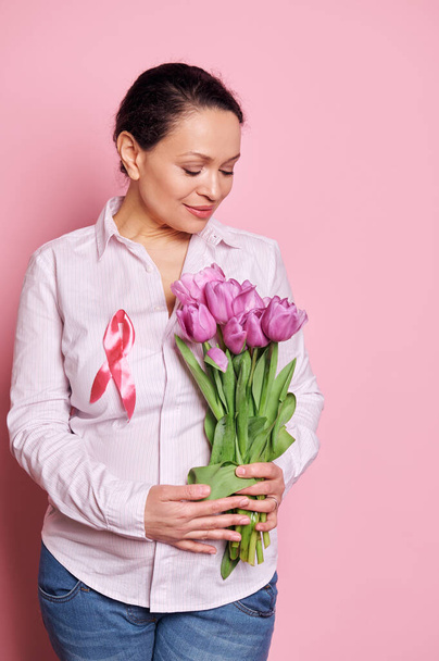 Pewny siebie portret na różowym tle ciężarnej ładnej kobiety, noszącej różową satynową wstążkę świadomości, trzymającej fioletowe tulipany na odizolowanym tle koloru. Różowy październik - miesiąc świadomości raka piersi - Zdjęcie, obraz