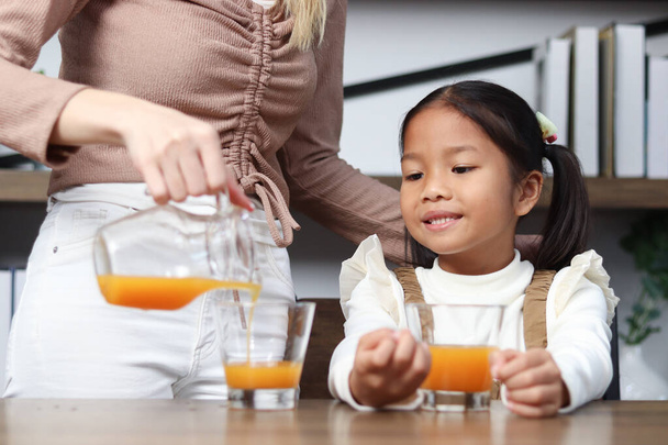 Asian happy family spending time together, mother pouring fresh orange juice from jug for her little cute daughter sitting in living room at home, young parent mom taking care kid to get healthy diet. - Foto, Bild